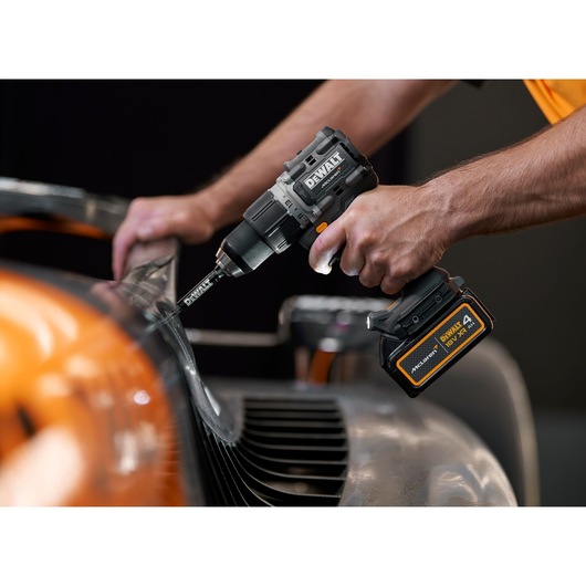A person uses a DEWALT cordless power drill to work on a metal surface, with an automotive part in the foreground.