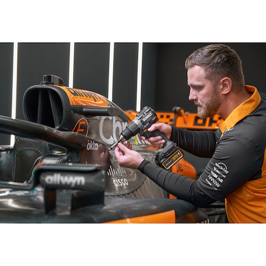 A person with a blurred face is using a DEWALT cordless drill to work on the side of an orange and black race car. The car