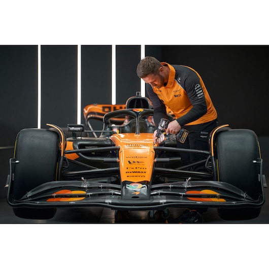 A person in an orange and black uniform is using a DEWALT power tool on the front section of an orange Formula 1 race car in