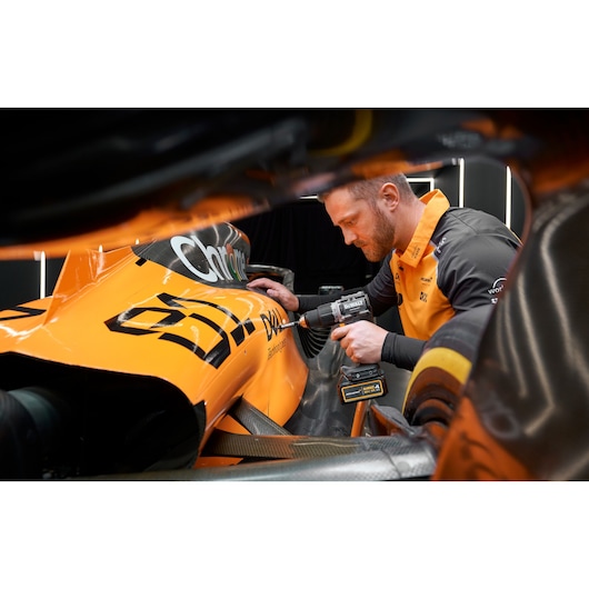A person in an orange and black outfit uses a DEWALT cordless drill on an orange race car. The focus is on the DEWALT