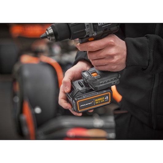 Person holding a DEWALT 18V cordless drill with McLaren branding, near an orange and black car.