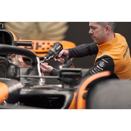 A person wearing a McLaren team jacket uses a DEWALT cordless drill on the front section of a Formula 1 car. The person's