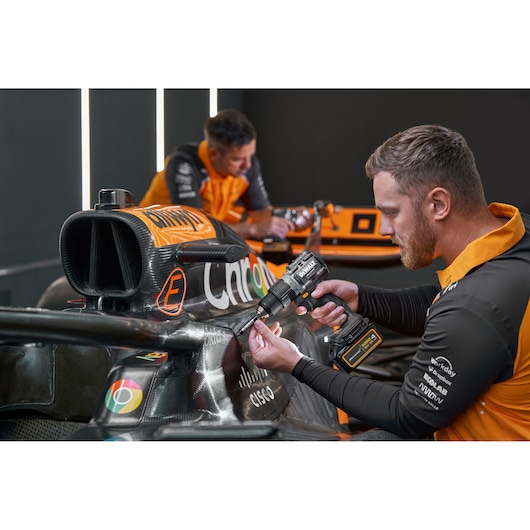 Two people wearing orange and black uniforms are working on a Formula 1 race car. One person is using a DEWALT cordless