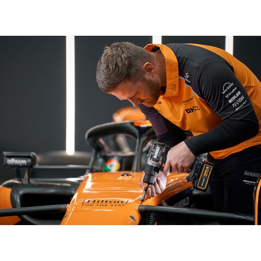 A person wearing an orange and black shirt uses a DEWALT cordless drill to work on the body of an orange Formula 1 race car