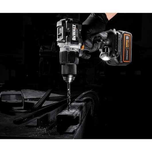 A DEWALT cordless drill is being used to drill into a metal surface in a workshop setting. The drill is equipped with a