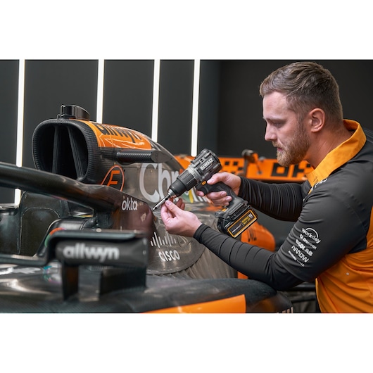 A person with a blurred face is using a DEWALT cordless drill to work on the carbon fiber body of a racing car in an indoor