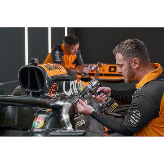 Two people with blurred faces are working on the body of an orange and black racing car using a DEWALT cordless power drill