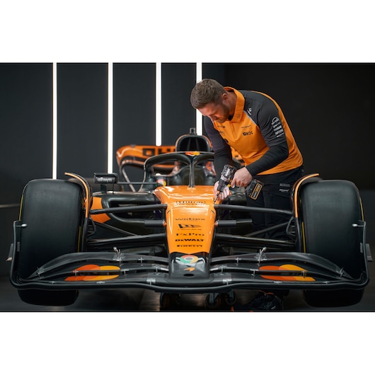 A person wearing an orange and black DEWALT-branded uniform is working on the front of an orange Formula 1 race car using a