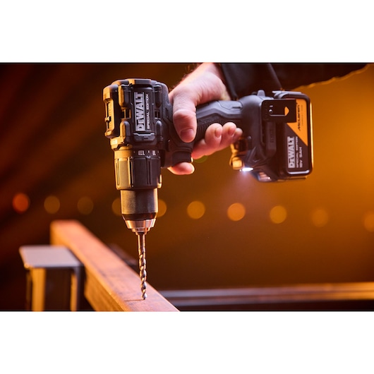 A close-up of a hand holding a DEWALT cordless drill with a drill bit attached, drilling into a metal surface. The drill is