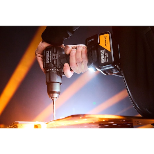 A close-up of a person using a DEWALT cordless drill to drill into a piece of metal, illuminated by bright orange lights in