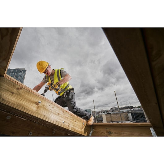 A construction worker wearing a yellow safety helmet and high-visibility vest uses a DEWALT cordless drill to work on a wooden beam at an outdoor building site, with cityscape visible in the background under a cloudy sky.