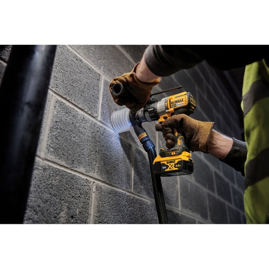 A person wearing work gloves uses a DEWALT cordless drill to bore a hole into a concrete block wall, with a dust extraction accessory attached for cleaner operation.