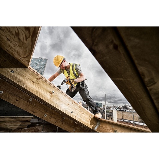 A construction worker wearing a yellow hard hat and safety vest is using a DEWALT cordless drill on a wooden beam at a building site, with city buildings visible in the background.
