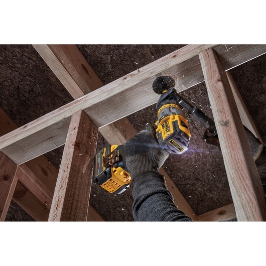 A person wearing gloves is using a DEWALT cordless drill to bore a hole in a wooden beam as part of a construction project. Wood shavings are flying from the drill.