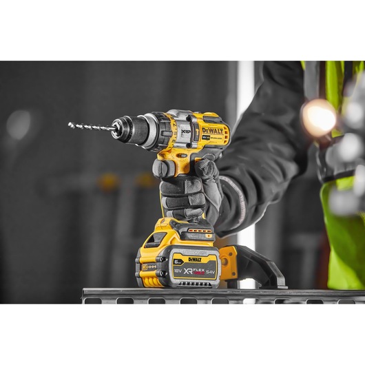 A close-up view of a person wearing gloves holding a yellow DEWALT DCD999NT cordless power drill with an attached battery and drill bit, positioned on a metal surface in a professional workshop setting.