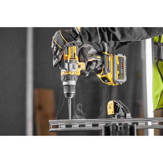A person wearing gloves is using a yellow and black DEWALT cordless power drill to drill into a metal beam. The drill is model DCD999NT and is equipped with a battery. The workspace appears industrial, and a clamp secures the metal beam.