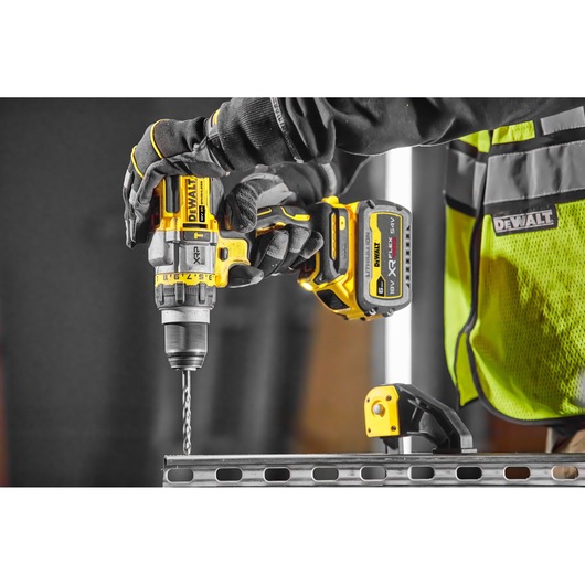 A person wearing gloves and a high-visibility vest uses a yellow and black DEWALT cordless drill to make a hole in a metal beam.