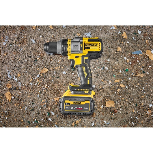 A yellow and black DEWALT cordless drill with an 18V XR FLEXVOLT 6Ah lithium-ion battery is placed on a rough, debris-covered concrete surface.