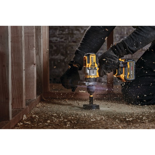 A person using a DEWALT cordless power drill to drill a hole in a wooden surface. Wood shavings are flying around as the drill is in use.