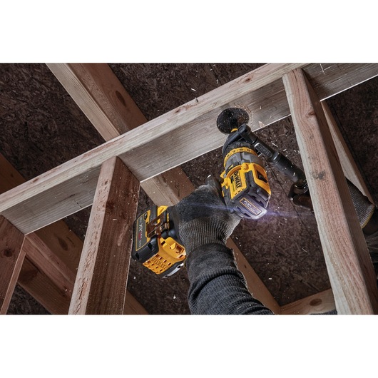 A person using a DEWALT cordless drill to bore a hole through a wooden beam in a construction setting. The user is wearing gloves and sawdust is visible as the drill operates.