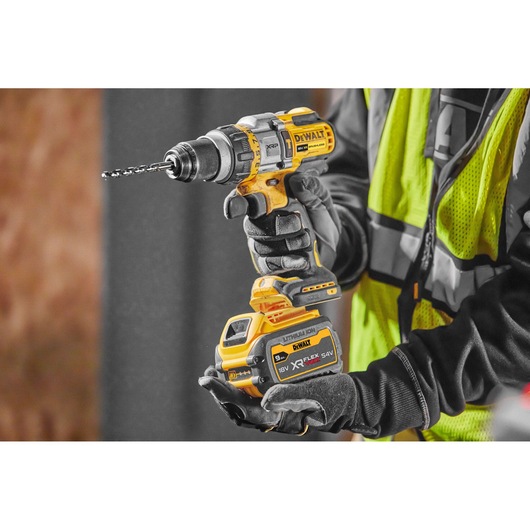 A person wearing work gloves and a safety vest is holding a DEWALT cordless power drill with a lithium-ion XR FLEXVOLT battery attached, preparing to use it.