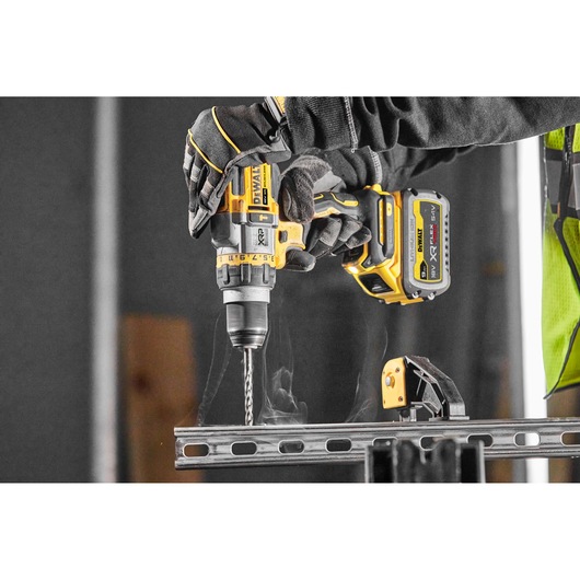 Close-up of a person wearing gloves using a DEWALT DCD999X1 cordless drill to drill through a metal beam, with visible smoke rising from the material.