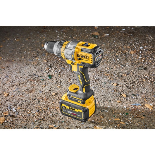 A DEWALT 18V XR FLEXVOLT cordless drill, model DCD999X1, standing upright on a rough, dusty surface. The drill is yellow and black with a large battery attached at the base.