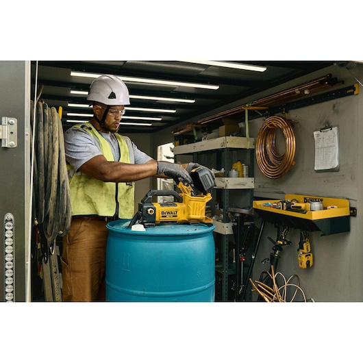 Worker using DEWALT DCE050N tool on blue barrel in a workshop, surrounded by cables, shelves, and equipment.