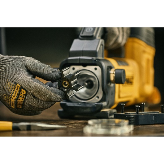 Gloved hand holding a part near a yellow DEWALT device. Tool and parts are on a workbench.