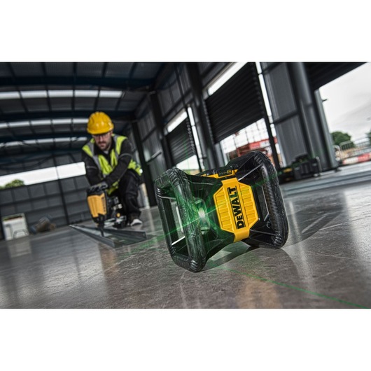 A DEWALT green laser level device is placed on a concrete floor inside a warehouse or construction site. The tool projects visible green lines, and a person wearing safety gear, including a helmet and high-visibility jacket, is working in the background.