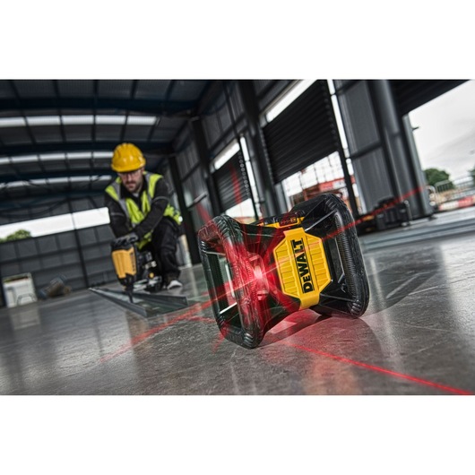 DEWALT laser level device emitting red laser lines on a construction site floor, with a worker in a yellow helmet and high-visibility vest working in the background.