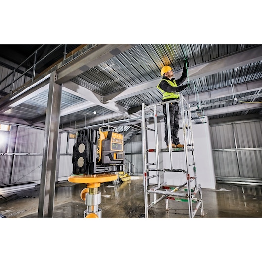 A DEWALT laser level device is set up on a tripod in an industrial building under construction. In the background, a worker wearing a hard hat and safety vest stands on scaffolding and marks or works on the metal ceiling.