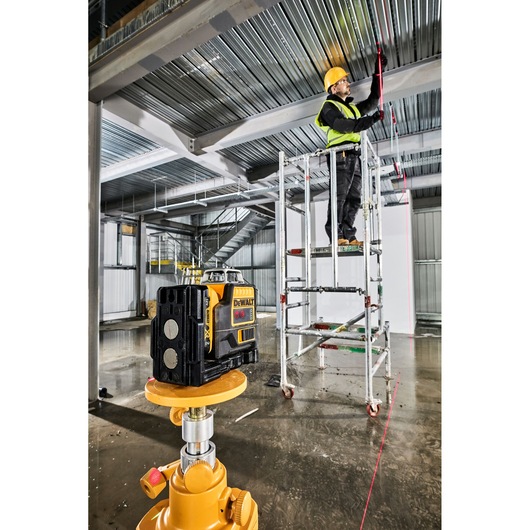 A DEWALT laser level device is set up on a tripod in the foreground of an industrial construction site. In the background, a worker wearing a yellow hard hat and high-visibility vest stands on scaffolding, marking a metal ceiling along the visible red laser line projected by the device. The area features metal beams and stairs, with construction materials visible throughout.