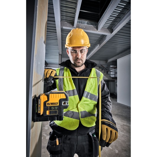 A person wearing a yellow hard hat, high-visibility vest, and work gloves is using a DEWALT DCE0822D1G green laser level tool attached to a metal column in an industrial or construction environment.