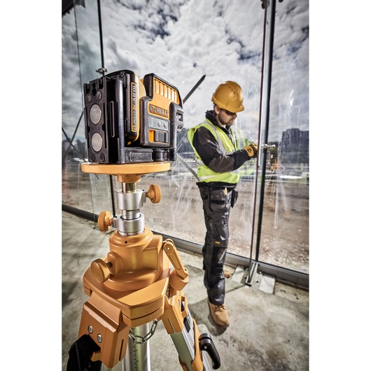 A DEWALT laser level device mounted on a tripod is shown in the foreground of a construction site. A construction worker, wearing a safety vest and hard hat, is working on a glass wall in the background.