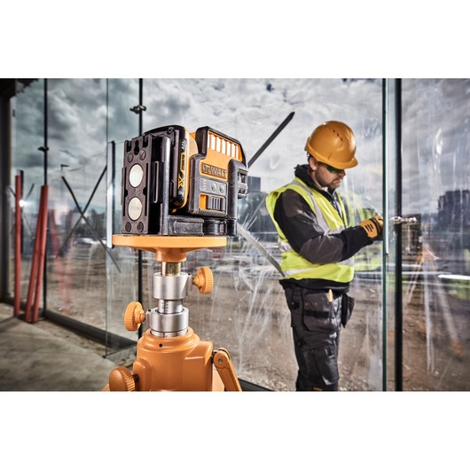 A DEWALT DCE0825D1G laser level tool mounted on a tripod is shown in the foreground of a construction site. In the background, a worker wearing a hard hat, high-visibility vest, and gloves is installing or inspecting glass panels.