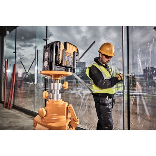 A DEWALT laser level device is mounted on a tripod at a construction site, with a person wearing a safety vest and hard hat working in the background near large glass panels.