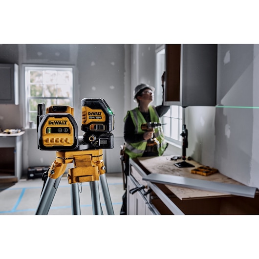 A DEWALT green laser level tool mounted on a tripod is shown in the foreground of a room under construction. The device projects a green laser line on the wall for alignment. In the background, a construction worker wearing a safety vest and helmet is using a power tool to install cabinetry.