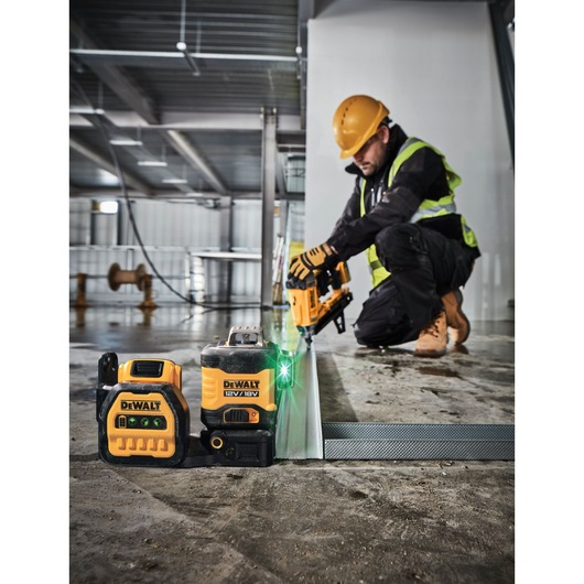 A worker wearing a yellow hard hat and safety vest is kneeling on a construction site floor, operating a Dewalt tool. In the foreground, a Dewalt 12V/18V green laser level and a Dewalt battery pack are placed on the floor, emitting a bright green laser line along a metal rail.