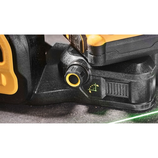 Close-up view of a DEWALT DCE089D1G18 laser level showing the adjustment knob and indicator symbol on the black and yellow casing. A green laser line is visible on the surface below.
