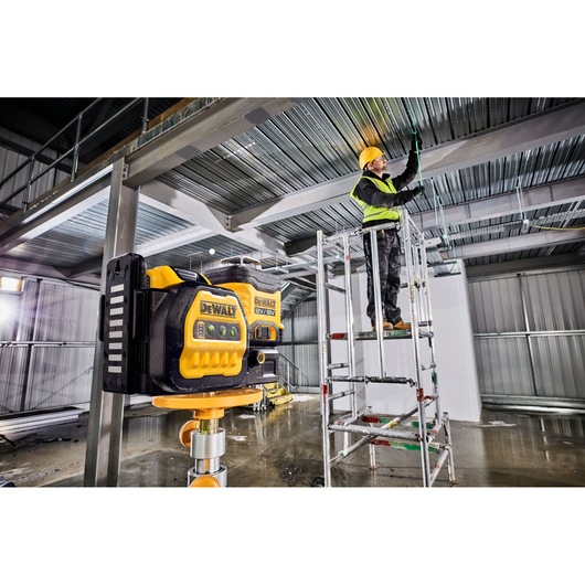A DEWALT DCE089NG18 laser level is mounted on a tripod inside an industrial building under construction. In the background, a worker wearing a hard hat and safety vest stands on a scaffold, installing wiring along the metal ceiling beams.
