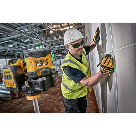 A DEWALT 12V/18V laser level is positioned on a tripod in the foreground. In the background, a construction worker wearing a safety helmet and reflective vest is marking a wall using the green laser line for alignment. The worker’s face is blurred for privacy. The scene takes place at a construction site with visible pipes and equipment.