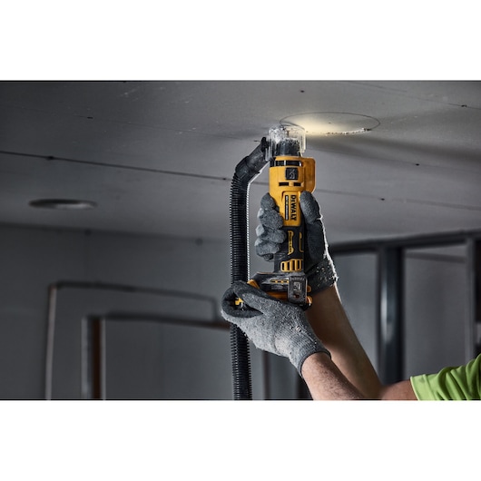 A person wearing gloves uses a yellow DEWALT cordless cutting tool to cut a circular hole in a ceiling panel. The tool is connected to a vacuum hose for dust extraction.