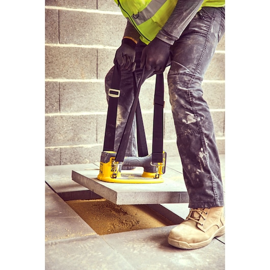 A person wearing work boots, gloves, and a high-visibility vest is using a DEWALT DCE590 suction lifter to lift and place a