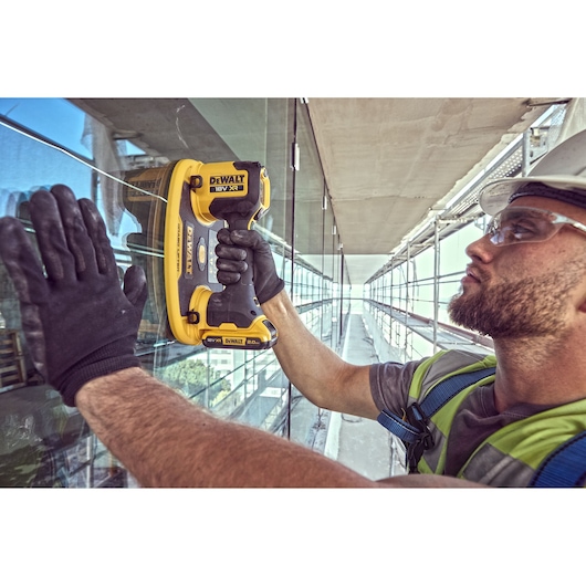 A person wearing safety gear uses a DEWALT DCE590 18V XR cordless tool to work on a glass surface, with scaffolding and a