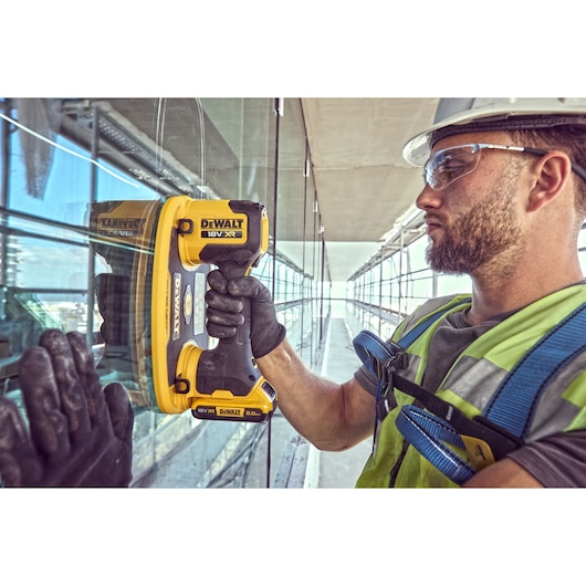 A person wearing safety gear uses a DEWALT DCE590 18V XR handheld power tool on a glass surface, likely for construction or