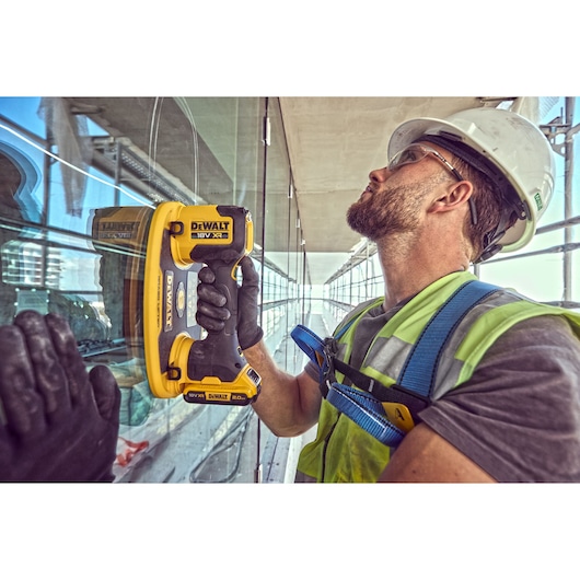 A construction worker wearing safety gear, including a helmet and gloves, uses a DEWALT 18V XR cordless tool to work on a