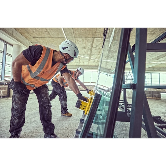 Two construction workers wearing safety helmets and orange high-visibility vests are handling a large sheet of glass using