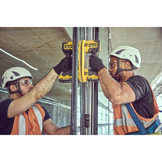 Two construction workers wearing safety helmets and vests use DEWALT DCE590 power tools to work on metal pipes inside a