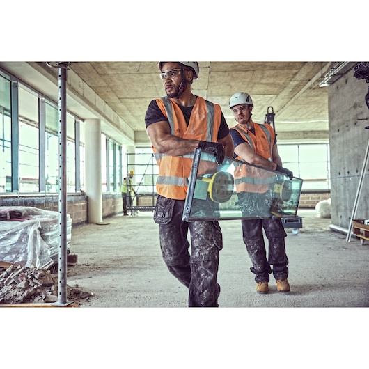 Two construction workers wearing orange safety vests and helmets are carrying a large glass panel with suction grips inside
